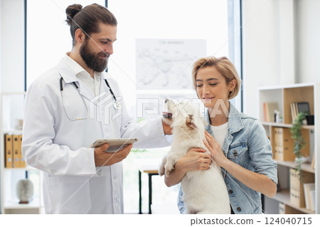 Man veterinarian with young adult woman owner examining white dog at modern clinic. Smiling doctor providing care. Concept of pet health, animal care, veterinary service, friendship. Man veterinarian with young adult woman owner examining white dog at modern clinic. Smiling doctor providing care. Concept of pet health, animal care, veterinary service, friendship. 124040715