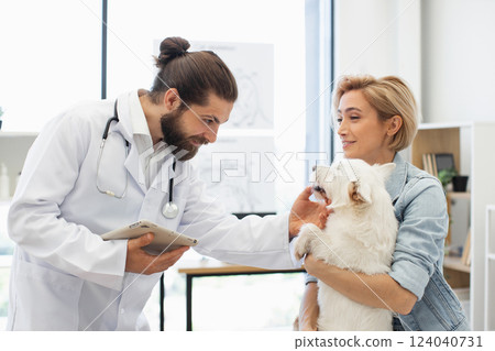 Man veterinarian examining small dog in light clinic setting while woman owner happily observes. Scene highlights animal care, trust, and professional pet healthcare service. Man veterinarian examining small dog in light clinic setting while woman owner happily observes. Scene highlights animal care, trust, and professional pet healthcare service. 124040731