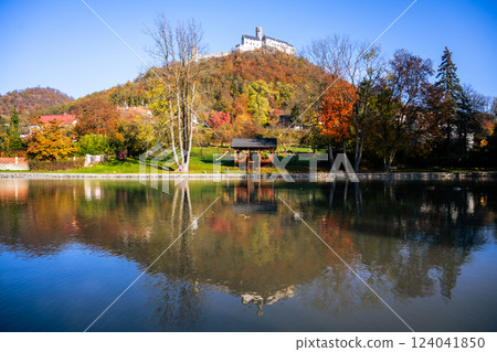 A serene autumn landscape featuring medieval Bezdez Castle atop a hill, surrounded by colorful foliage and mirrored in the calm waters below, showcasing the beauty of the Czech countryside. 124041850