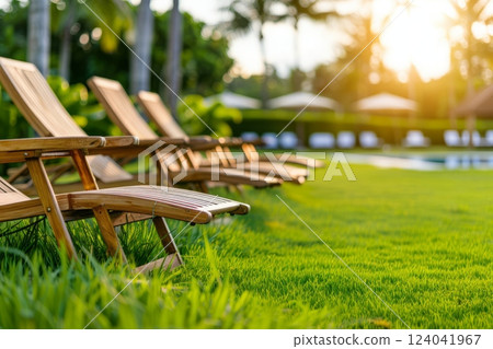 Wooden garden loungers placed on green grass near a pool, surrounded by lush tropical plants. 124041967