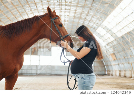 Side view. Beautiful pregnant woman in casual clothes is with the horse on a stable 124042349