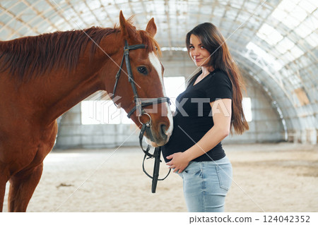 Looking at the camera. Beautiful pregnant woman in casual clothes is with the horse on a stable Looking at the camera. Beautiful pregnant woman in casual clothes is with the horse on a stable 124042352
