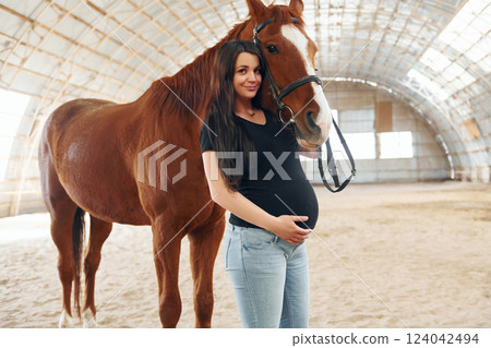 Looking at the camera. Beautiful pregnant woman in casual clothes is with the horse on a stable Looking at the camera. Beautiful pregnant woman in casual clothes is with the horse on a stable 124042494