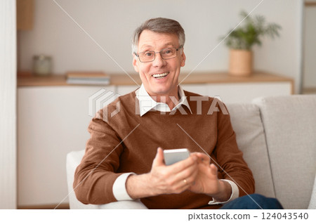 Mobile App. Happy Elderly Man Using Smatphone Smiling At Camera Sitting On Sofa Indoor. 124043540