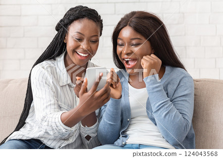 Good News. Happy Girlfriends Looking At Smartphone Screen With Excitement, Sitting On Couch At Home Together. Good News. Happy Girlfriends Looking At Smartphone Screen With Excitement, Sitting On Couch At Home Together. 124043568