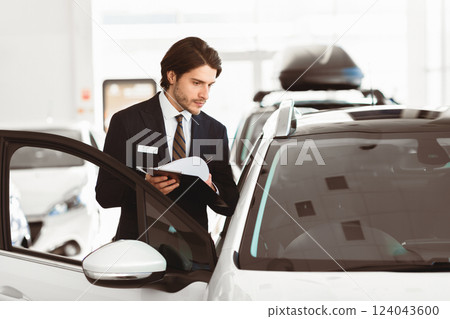 Vehicle Dealership. Car salesperson with clipboard checking auto specifications in local dealership showroom Vehicle Dealership. Car salesperson with clipboard checking auto specifications in local dealership showroom 124043600