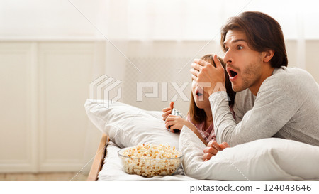 Young Dad And His Little Girl Are Watching Horror, laying on bed and eating popcorn, father closing daughter eyes, copy space 124043646