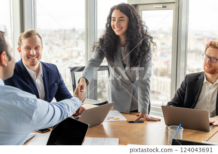 A businesswoman in a gray blazer extends her hand to shake hands with a male colleague in a business meeting. The group is sitting around a wooden conference table in front of large windows A businesswoman in a gray blazer extends her hand to shake hands with a male colleague in a business meeting. The group is sitting around a wooden conference table in front of large windows 124043691