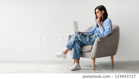Full length of serious young woman looking at laptop screen, sitting in armchair, having problem with online work or studies against white studio wall, banner with copy space Full length of serious young woman looking at laptop screen, sitting in armchair, having problem with online work or studies against white studio wall, banner with copy space 124043727