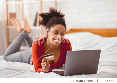 Online Shopping Concept. Cheerful Black Lady Using Laptop And Credit Card Purchasing Things On Internet Lying In Bed Indoor 124043728
