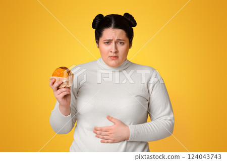 Negative Effects of Fast Food Concept. Plump Girl Holding Burger And Touching Abdomen, Having Stomach Ache, Yellow Studio Background, Copy Space 124043743