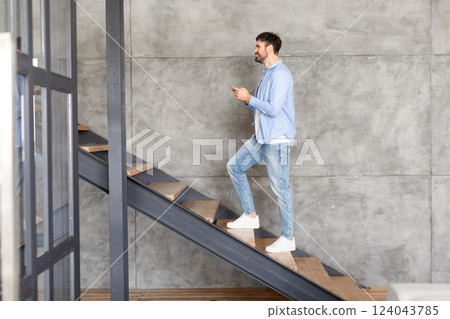 This image shows a man walking up a modern staircase in a home. The man is casually dressed in a blue shirt and jeans and is holding a phone in his hand. The stairs are made of wood This image shows a man walking up a modern staircase in a home. The man is casually dressed in a blue shirt and jeans and is holding a phone in his hand. The stairs are made of wood 124043785