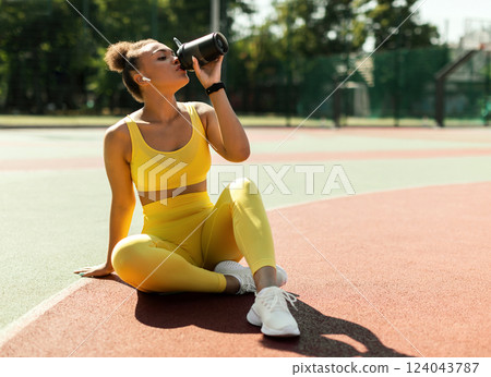 Energy, Vitality, Wellbeing Concept. Fit African American woman in wireless earbuds sitting on basketball field ground, holding shaker, drinking water or protein, taking break after outdoors exercise Energy, Vitality, Wellbeing Concept. Fit African American woman in wireless earbuds sitting on basketball field ground, holding shaker, drinking water or protein, taking break after outdoors exercise 124043787
