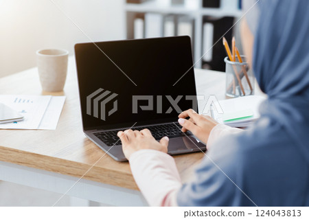Unrecognizable muslim woman working on laptop with black screen, typing on keyboard, sitting in modern office, over shoulder view, mockup 124043813