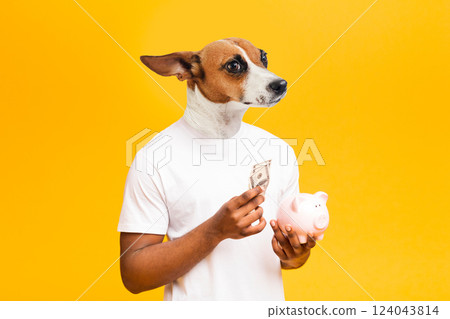 A man with the head of a dog holds a piggy bank and some cash in his hands while looking to the left. He is wearing a white t-shirt and standing in front of a bright yellow background. 124043814