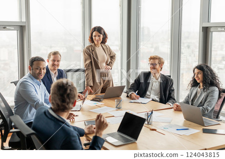 A group of diverse business professionals are seated around a large conference table in a modern office space. One woman stands at the head of the table, while the others focus on the discussion A group of diverse business professionals are seated around a large conference table in a modern office space. One woman stands at the head of the table, while the others focus on the discussion 124043815
