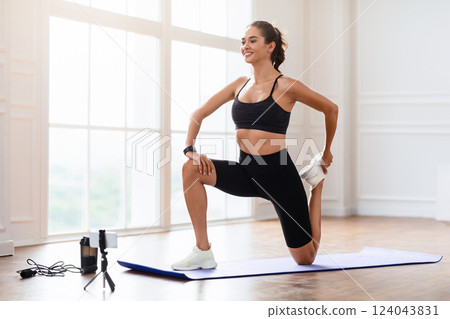 Smiling Sporty Woman Training At Home In Front Of Smartphone On Tripod, Beautiful Female Fitness Blogger Shooting Online Video Fitness Tutorials, Showing Stretching Legs Exercises At Camera Smiling Sporty Woman Training At Home In Front Of Smartphone On Tripod, Beautiful Female Fitness Blogger Shooting Online Video Fitness Tutorials, Showing Stretching Legs Exercises At Camera 124043831