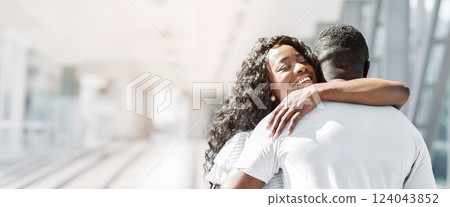 Black woman with long, curly black hair is hugging a man from behind. She is smiling broadly, looking over her shoulder at the viewer. He is wearing a white shirt, copy space 124043852
