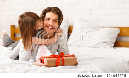 Cute little girl greeting her happy daddy with Father Day, giving him gift box and greeting card, hugging and kissing, laying on bed together at home, free space Cute little girl greeting her happy daddy with Father Day, giving him gift box and greeting card, hugging and kissing, laying on bed together at home, free space 124043856