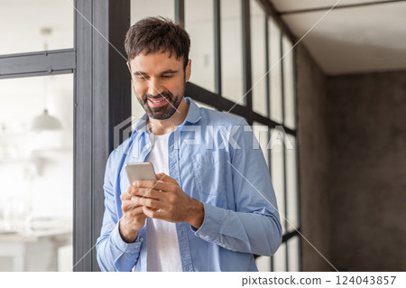 A man with a beard stands near a window in a modern office setting, smiling as he uses his smartphone. His blue shirt and white undershirt are casual, copy space 124043857