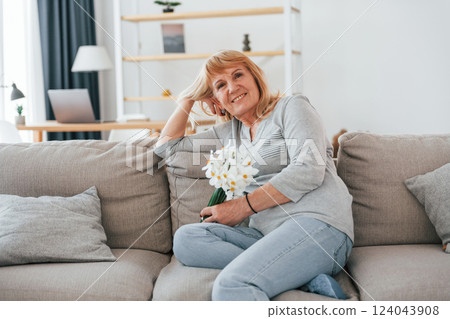 Sitting on a sofa with flowers in hands. Senior woman with red hair is at home alone 124043908