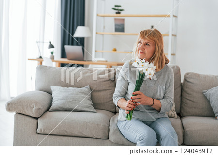 Sitting on a sofa with flowers in hands. Senior woman with red hair is at home alone Sitting on a sofa with flowers in hands. Senior woman with red hair is at home alone 124043912
