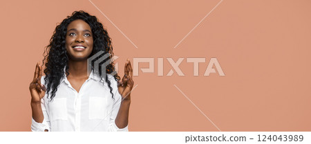 A young black woman with long, curly black hair is standing against a peach-colored background. She is smiling, and her fingers are crossed as she looks up with hopeful anticipation. A young black woman with long, curly black hair is standing against a peach-colored background. She is smiling, and her fingers are crossed as she looks up with hopeful anticipation. 124043989