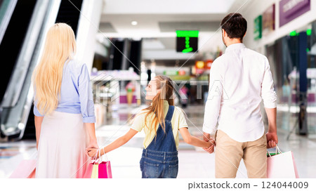 A family of three walks through a shopping mall. They are holding hands, and the parents each carry a shopping bag. The young girl is looking back at something behind them 124044009