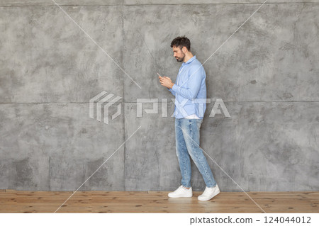A man in a light blue shirt and blue jeans walks past a concrete wall while looking at his smartphone. He is wearing white sneakers. The floor is wooden, copy space 124044012