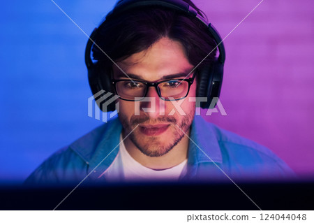 A young man is seen in close-up, wearing headphones and glasses, focused on a computer screen in front of him. The room is lit in blue and purple lighting 124044048