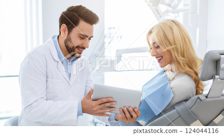 Smiling man dentist showing female patient jaw x-ray on digital tablet, dental clinic interior. Bearded man stomatologist having conversation with lady in dental chair, holding pad, side view 124044116