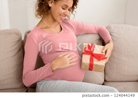 Baby shower gift. Black pregnant woman holding gift box, resting on sofa 124044149