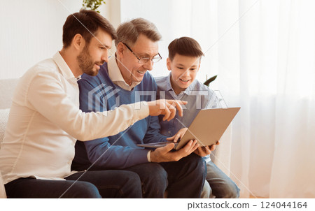 Grandfather Learning To Use Laptop Computer With Adult Son And Grandson Sitting On Sofa Indoor. Selective Focus Grandfather Learning To Use Laptop Computer With Adult Son And Grandson Sitting On Sofa Indoor. Selective Focus 124044164