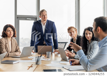 A group of business professionals are gathered around a large wooden table in a modern office space. The men and women are engaged in a meeting, discussing ideas and strategies 124044255