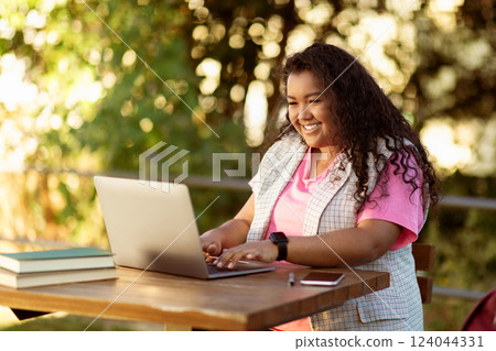A woman with curly hair smiles while using her laptop at a wooden table surrounded by greenery. She has books and a phone nearby, enjoying a pleasant outdoor workspace in the afternoon. A woman with curly hair smiles while using her laptop at a wooden table surrounded by greenery. She has books and a phone nearby, enjoying a pleasant outdoor workspace in the afternoon. 124044331