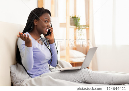 Phone Call. Excited Black Girl Talking On Cellphone Using Laptop Sitting In Bed At Home. Selective Focus, Copy Space 124044421