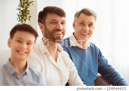 Happy Elderly Man With Middle-Aged Son And Grandson Smiling At Camera Sitting On Couch At Home. Selective Focus Happy Elderly Man With Middle-Aged Son And Grandson Smiling At Camera Sitting On Couch At Home. Selective Focus 124044423