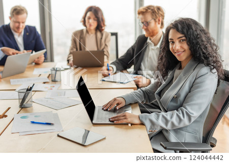 A woman with curly hair sits at a conference table and types on her laptop while other business professionals work around her. A large window with a city view is visible in the background. 124044442