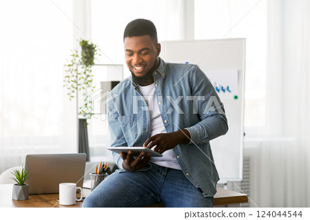 Modern technologies for work. Young african american ceo using digital tablet in office, sitting on table and smiling 124044544