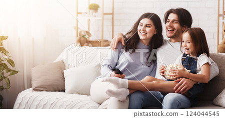Family Bonding. Happy parents and their little daughter watching tv at home together, eating popcorn and relaxing on couch in living room 124044545