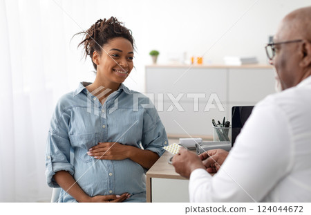 Pregnancy, Obstetrics, Gynaecology, Medicine, Healthcare And People Concept. Black male gynecologist giving pills to smiling pregnant woman during appointment at hospital. Visit To A Doctor Concept Pregnancy, Obstetrics, Gynaecology, Medicine, Healthcare And People Concept. Black male gynecologist giving pills to smiling pregnant woman during appointment at hospital. Visit To A Doctor Concept 124044672