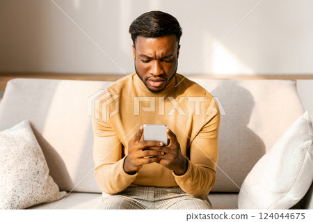 Discontended African American Man Holding Smaprtphone And Frowning Reading Message Sitting On Sofa Indoor. Selective Focus Discontended African American Man Holding Smaprtphone And Frowning Reading Message Sitting On Sofa Indoor. Selective Focus 124044675