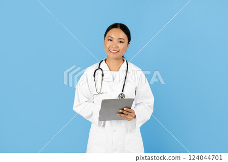 A healthcare professional Asian lady stands confidently, wearing a white coat and stethoscope, while holding a tablet in front of a light blue background, ready to assist patients. 124044701