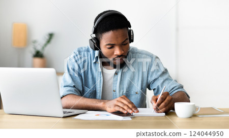 Concentrated african american guy in headphones studying foreign language online through video conference and making notes in notebook, panorama with copy space 124044950