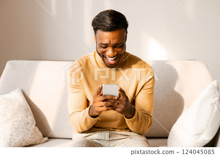 Mobile Gaming. Excited Afro Guy Using Cellphone Playing Games Sitting On Couch At Home. Mobile Gaming. Excited Afro Guy Using Cellphone Playing Games Sitting On Couch At Home. 124045085