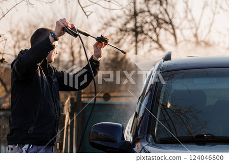 Man Washing Black Car with High-Pressure Water Jet in Outdoor Setting at Sunset 124046800