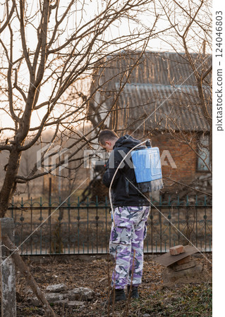 Man Spraying Garden Trees with Backpack Sprayer in Early Spring Yard Maintenance 124046803