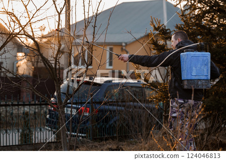 Man Spraying Trees in Garden with Backpack Sprayer During Sunset in Residential Area Man Spraying Trees in Garden with Backpack Sprayer During Sunset in Residential Area 124046813
