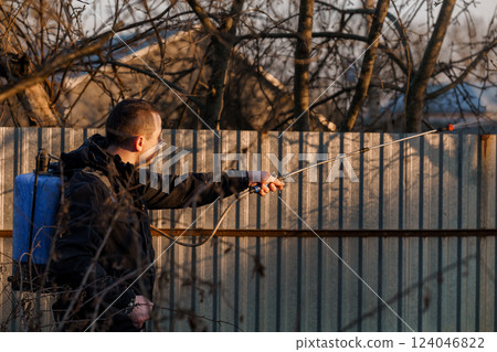 Man Holding Sprayer Spraying Chemicals onto Trees in Backyard During Sunset 124046822