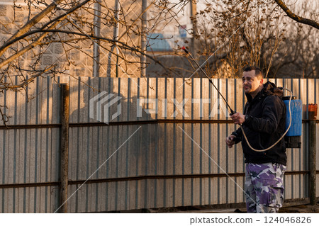 Man in Purple Camouflage Pants Spraying Chemicals in Backyard During Evening Sunset with Silhouetted Fence and Leafless Trees 124046826